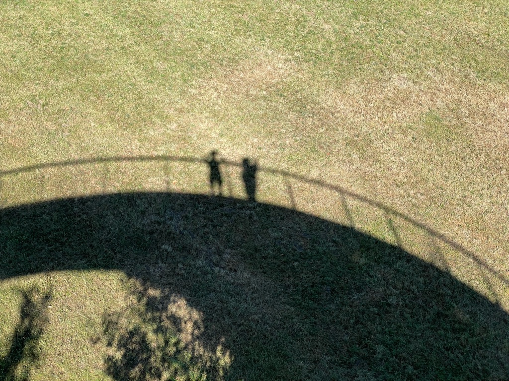 Shadows of two people are seen standing on a curved structure, likely a bridge or railing, casting onto a grassy surface. The angle of light creates a distinct silhouette effect.