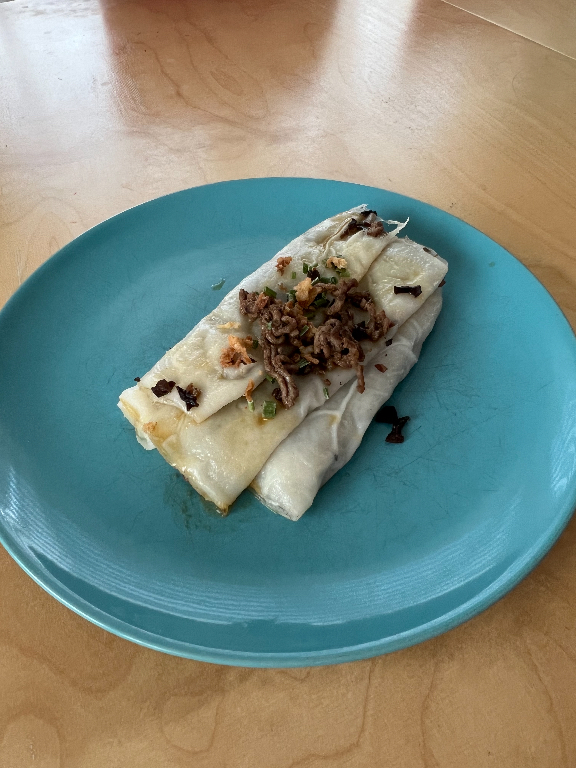 A dish of fresh rice wraps is neatly folded on a light blue plate. The Banh Cuons are topped with a mixture of sautéed ground meat, chopped green onions, and fried shallots, adding a touch of color and texture. The plate is set on a wooden table.