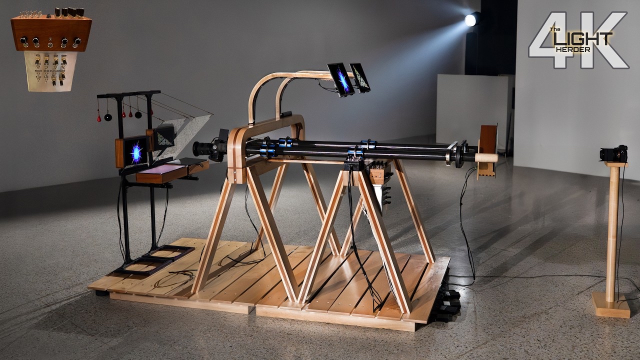 The image shows a YouTube video thumbnail featuring an intricate, handmade light contraption composed of wooden stands and metal rods. The device appears to be a complex setup for light manipulation, with cables connecting various components, including small screens and reflective surfaces. At the top right, the text “The Light Herder 4K” is displayed. The scene is brightly lit, highlighting the wooden framework and the detailed apparatus on a polished, light-colored floor.