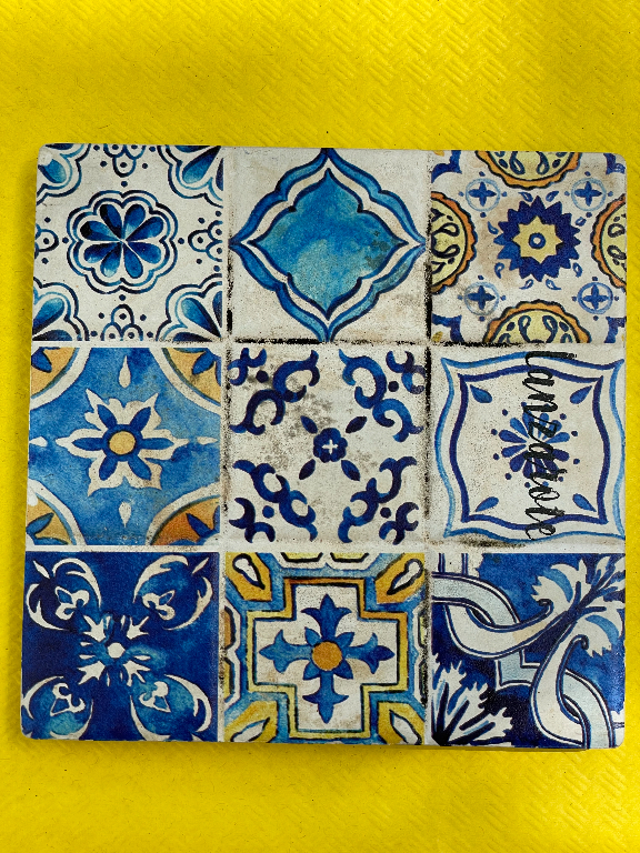 A square arrangement of nine decorative ceramic tiles, each with intricate blue and yellow designs, is laid out against a textured yellow background. The patterns on the tiles include floral and geometric motifs with varying shades of blue and accents of white and yellow.