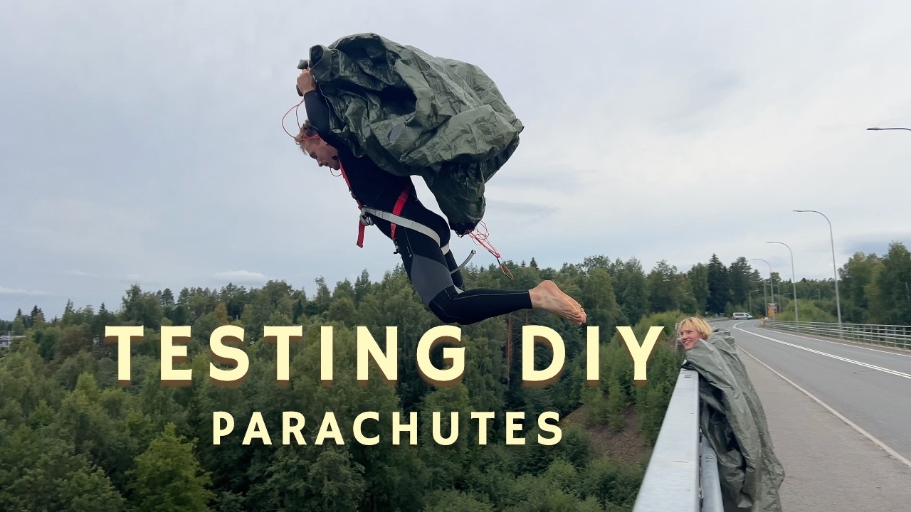 A person wearing a black wetsuit is jumping off a bridge while holding a DIY parachute made from a tarp. Another person looks on from the bridge, partially wrapped in a similar tarp. The text &ldquo;TESTING DIY PARACHUTES&rdquo; is overlaid on the image. The backdrop shows greenery and trees under a cloudy sky.