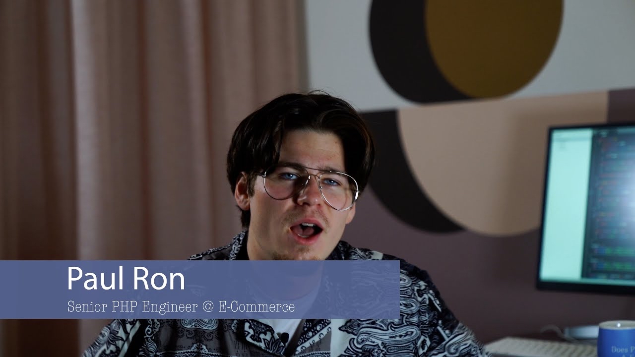 The image is a YouTube video thumbnail featuring a person with glasses and dark hair, sitting indoors. They are speaking or expressing something. The background includes a patterned wall and a computer screen. A text overlay in the lower-left corner reads: “Paul Ron, Senior PHP Engineer @ E-Commerce.”